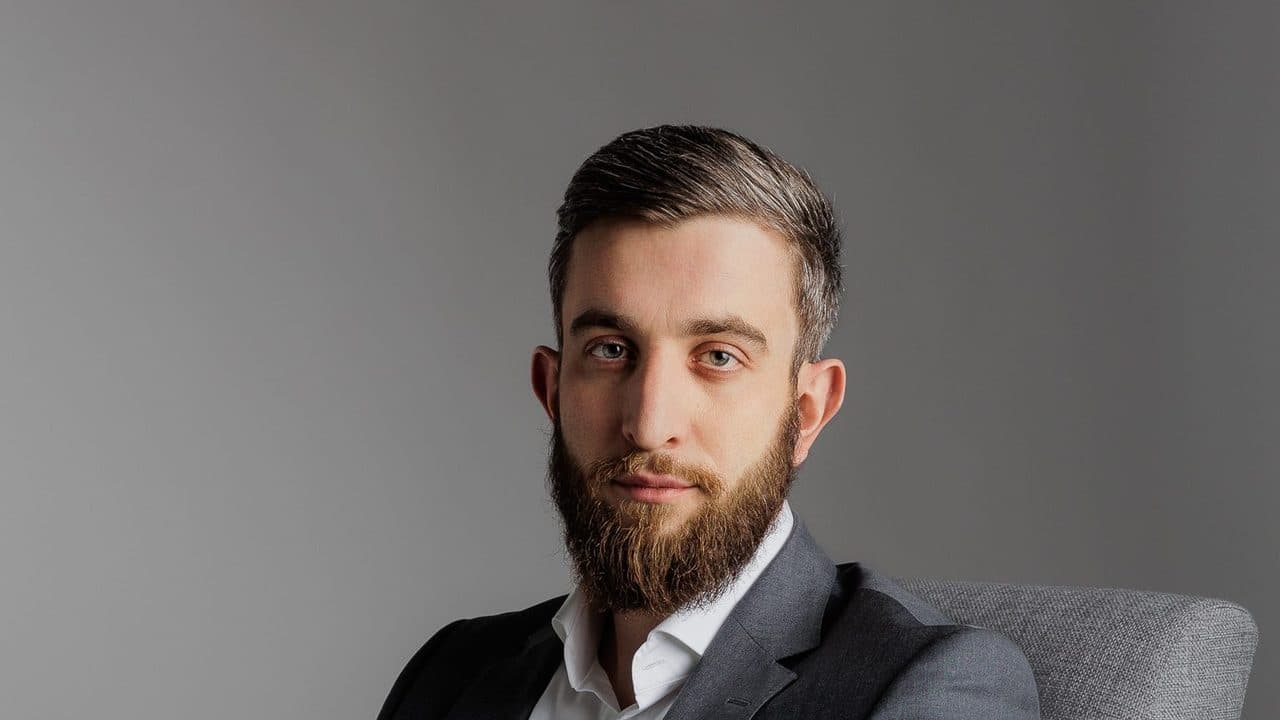 Studyjny portret brodatego mężczyzny w marynarce siedzącego w fotelu na szarym tle
